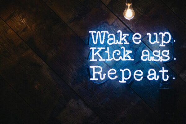 Neon sign on a dark wooden wall displaying a motivational message with two exposed light bulbs hanging above.