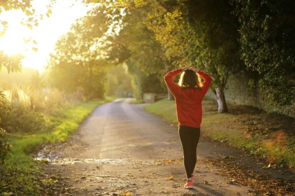 Woman walking on a sunlit path through a forest in the early morning, symbolizing healthy morning routines and active aging.