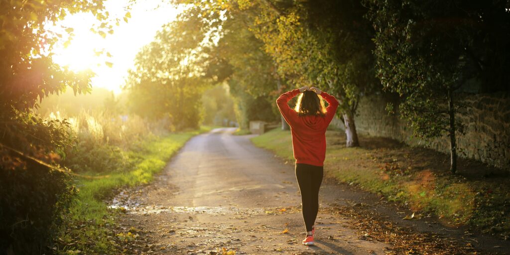 Woman walking on a sunlit path through a forest in the early morning, symbolizing healthy morning routines and active aging.