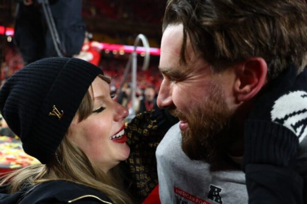 Travis Kelce and Taylor Swift smiling and embracing on the field after a football game, surrounded by a cheering crowd.