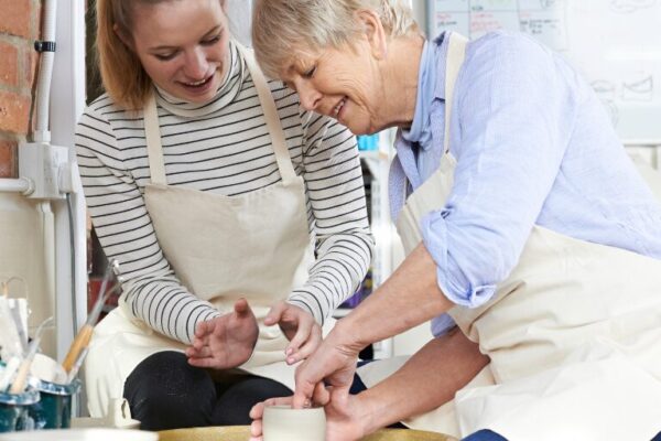 A mother and daughter wearing aprons shape clay together on a pottery wheel in a bright art studio.