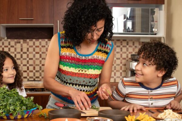 When Is Mexican Mother’s Day 2025? What to Know About Día de las Madres A woman cooks with two children in a cozy kitchen.