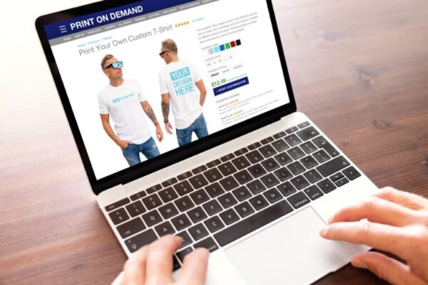 Person browsing a print-on-demand website on a laptop, customizing a white t-shirt with the text “Your Design Here” for an online store.
