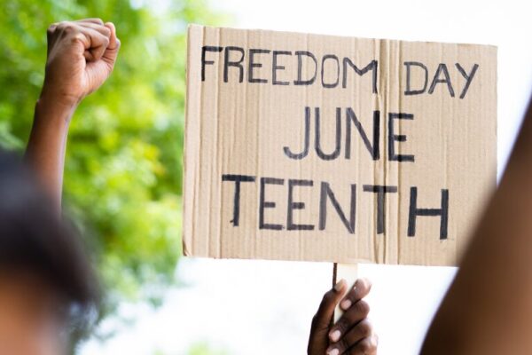 A cardboard sign reading "Freedom Day Juneteenth" is held up during a demonstration, with raised fists visible in the background, symbolizing unity and celebration.