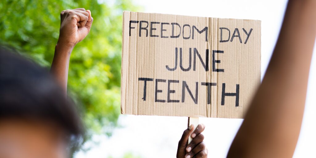 A cardboard sign reading "Freedom Day Juneteenth" is held up during a demonstration, with raised fists visible in the background, symbolizing unity and celebration.