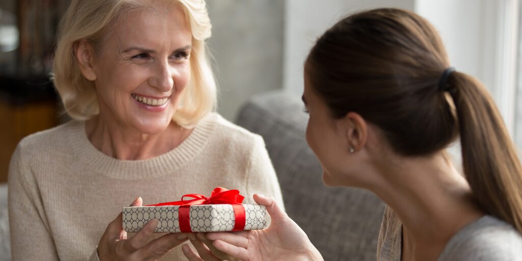 Daughter giving a smiling older woman a gift wrapped with a red ribbon, capturing a heartfelt Mother's Day moment.