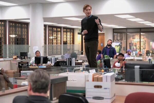 Man standing on a desk addressing coworkers in a busy office setting from the TV show "The Paper"