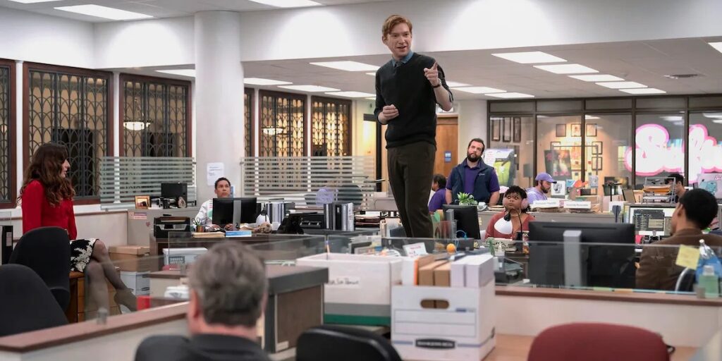 Man standing on a desk addressing coworkers in a busy office setting from the TV show "The Paper"