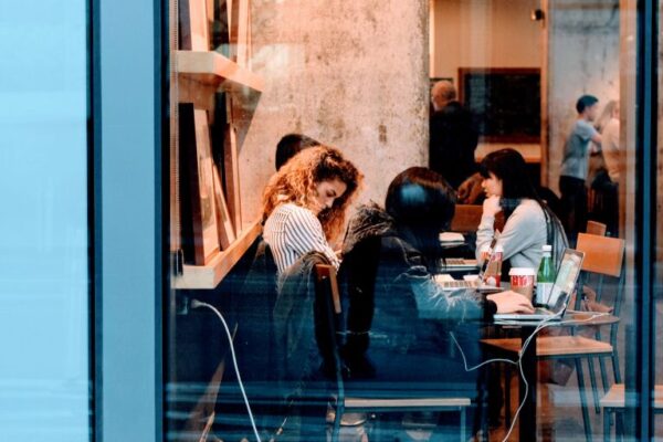 People working and socializing inside a busy coffee shop with laptops and drinks, viewed through a large window from the street