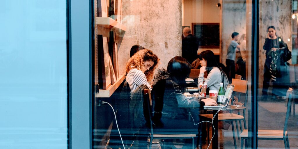 People working and socializing inside a busy coffee shop with laptops and drinks, viewed through a large window from the street