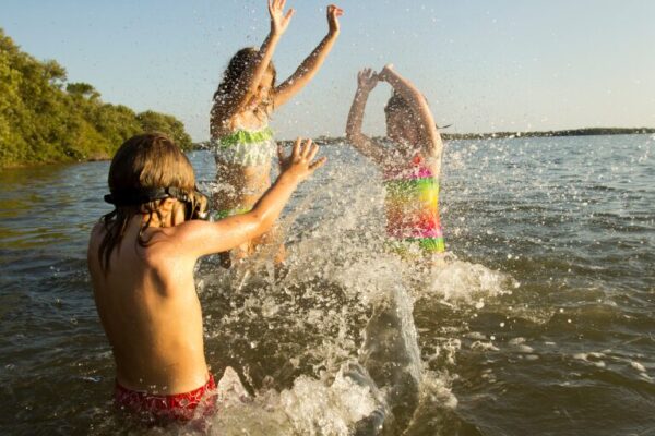 Top Family Lakeside Destinations for Memorial Day Weekend 2025 Three children in colorful swimsuits joyfully splashing water as they play together in a sunlit lake, with green trees lining the distant shore.