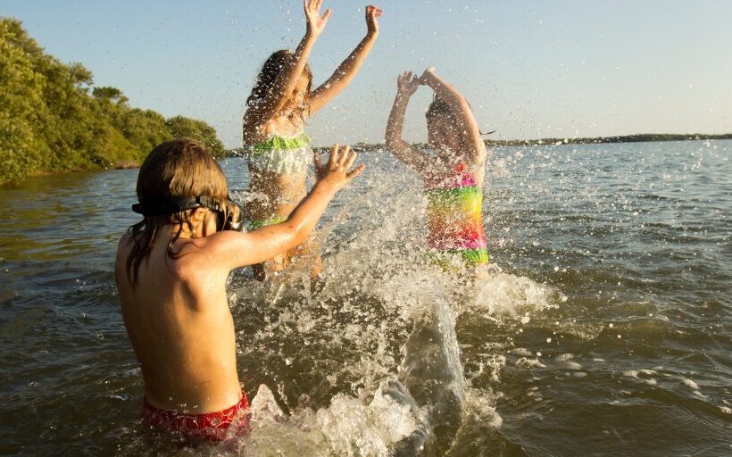 Top Family Lakeside Destinations for Memorial Day Weekend 2025 Three children in colorful swimsuits joyfully splashing water as they play together in a sunlit lake, with green trees lining the distant shore.