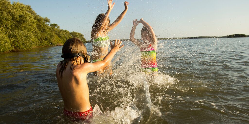 Three children in colorful swimsuits joyfully splashing water as they play together in a sunlit lake, with green trees lining the distant shore.